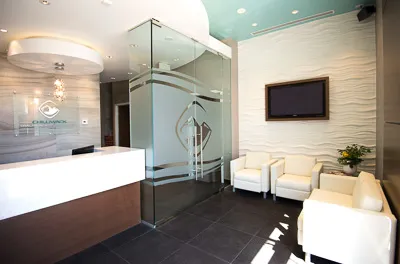 Reception desk and waiting area with white leather armchairs, a wall-mounted TV, and frosted glass walls.