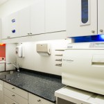 Clean dental sterilization room featuring white cabinetry, a dark countertop sink, and an autoclave machine.